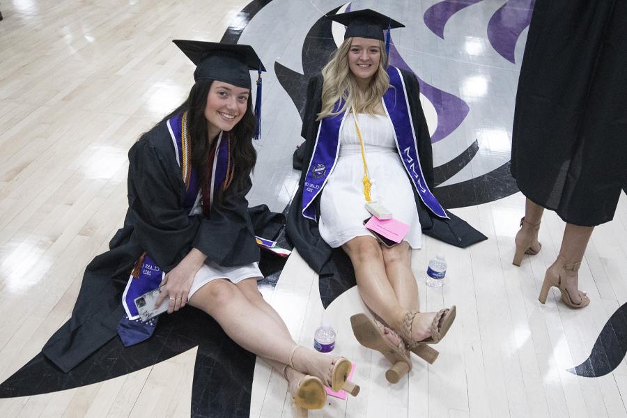 Two students wear academic regalia and sit on the gym floor by a large Warhawk head.